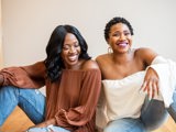 two black women sitting with arms on knees laughing